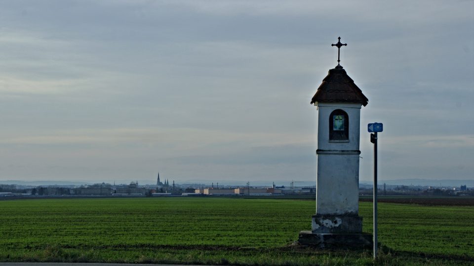 Boží muka na kraji obce a v pozadí Olomouc